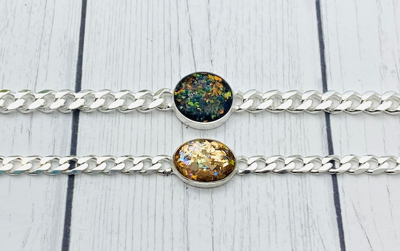 May include: Two silver-toned chain bracelets featuring central oval settings. The top bracelet contains a dark, iridescent, multi-colored flecked inlay, while the bottom bracelet features a gold-toned, shimmering, metallic-flecked inlay.