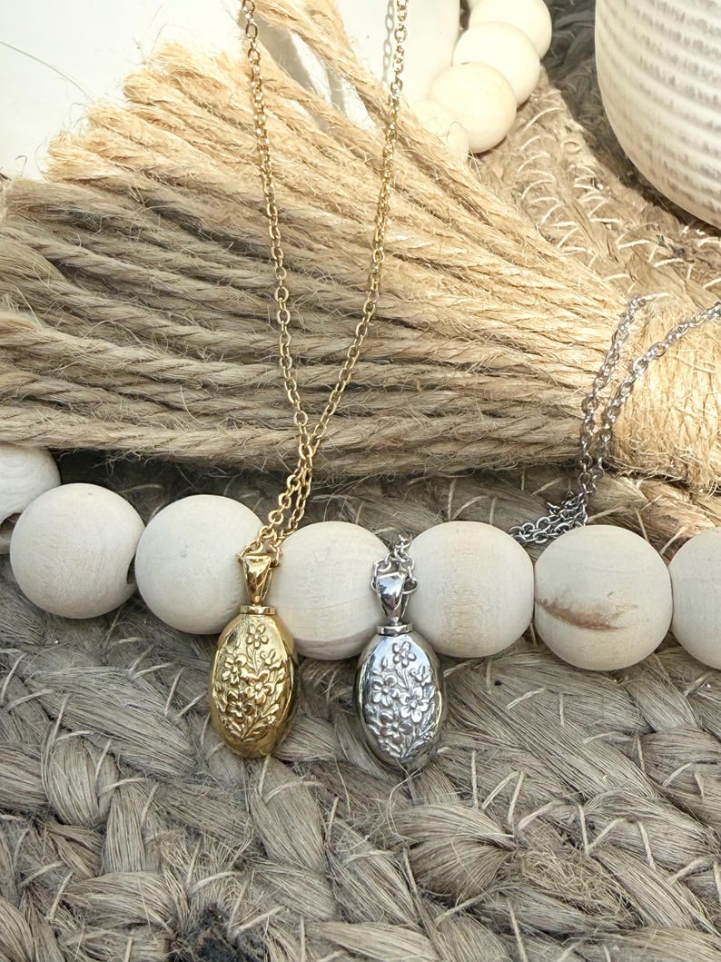 May include: Two necklaces with oval pendants. One pendant is gold-toned with a floral design, and the other is silver-toned with a similar floral pattern. Both pendants hang from delicate chains. The necklaces rest on a woven surface.