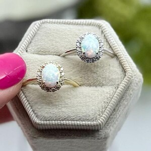 May include: Two opal rings with halo settings are displayed in a gray velvet ring box. One ring has a gold band, the other a silver band. The opals have a pearlescent sheen, and the halo settings are encrusted with small, clear stones.