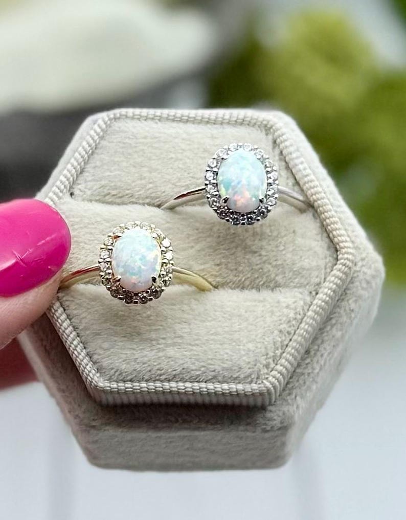 May include: Two opal rings with halo settings are displayed in a gray velvet ring box. One ring has a gold band, the other a silver band. The opals have a pearlescent sheen, and the halo settings are encrusted with small, clear stones.
