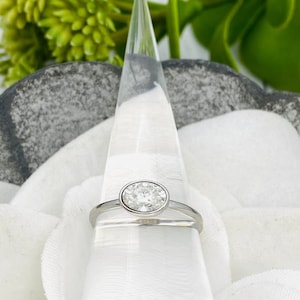 May include: A silver ring with a round, clear gemstone set in a bezel setting. The ring is displayed on a white ring holder.