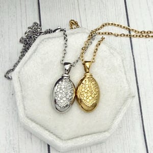May include: Two oval-shaped pendant necklaces. One is silver-toned, the other gold-toned. Both feature a floral design and hang on delicate chains. The pendants rest on a white, hexagonal surface against a light wood background.