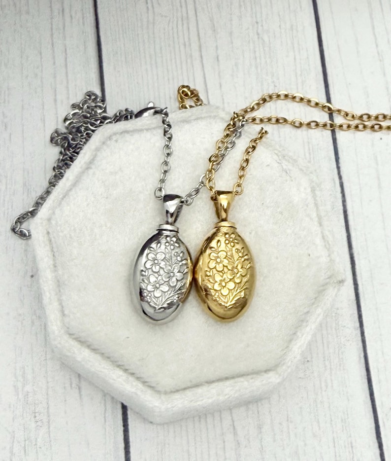 May include: Two oval-shaped pendant necklaces. One is silver-toned, the other gold-toned. Both feature a floral design and hang on delicate chains. The pendants rest on a white, hexagonal surface against a light wood background.