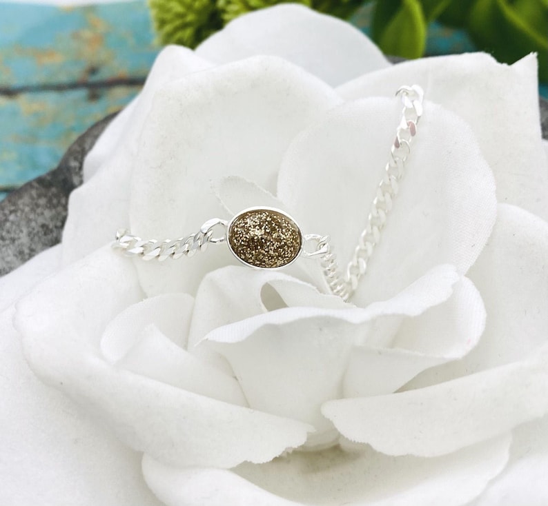 May include: A silver-toned chain bracelet featuring a central oval-shaped charm with a textured, shimmering gold-colored surface. The bracelet is composed of small, interlocking metal links that connect to the central oval piece on both sides.