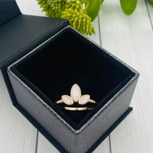 K&ouml;nnte beinhalten: Ein goldener Ring mit drei wei&szlig;en, ovalen Steinen, die in einem kronenartigen Design gefasst sind. Der Ring befindet sich in einer schwarzen Samt-Ringbox.