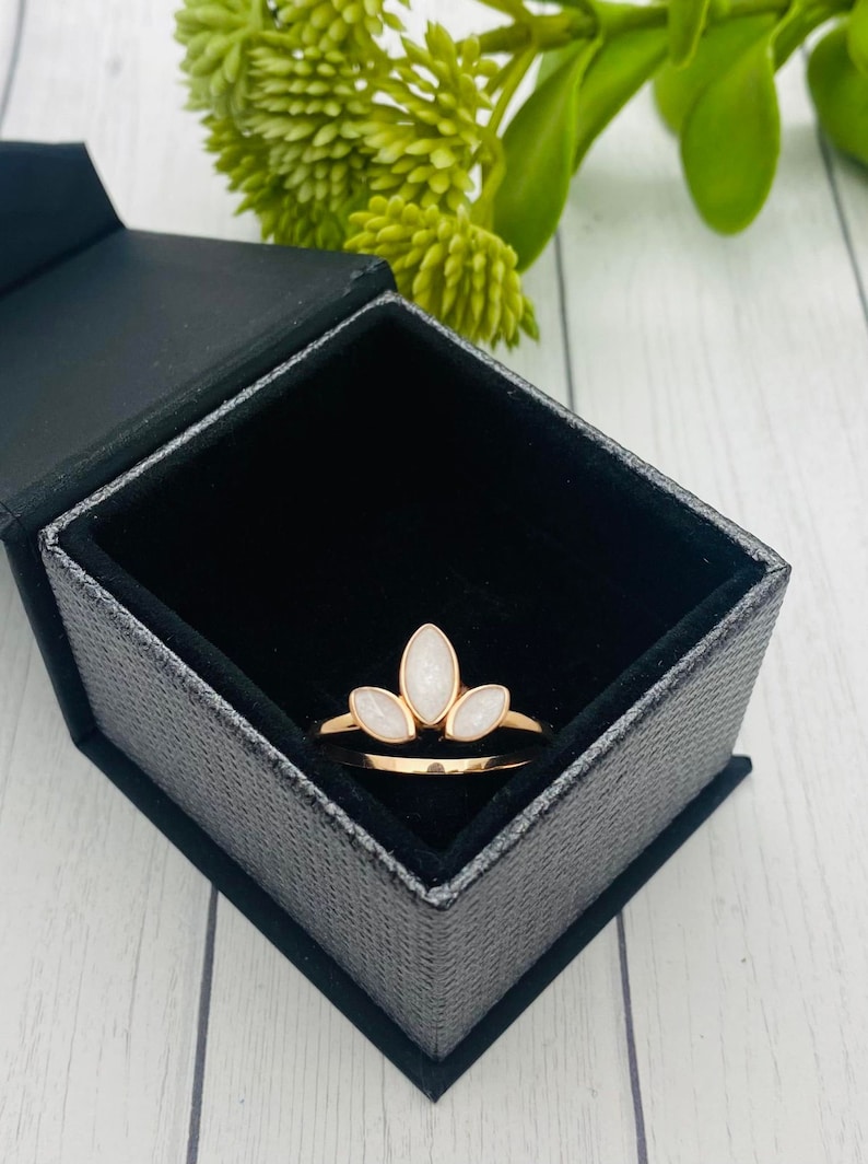 K&ouml;nnte beinhalten: Ein goldener Ring mit drei wei&szlig;en, ovalen Steinen, die in einem kronenartigen Design gefasst sind. Der Ring befindet sich in einer schwarzen Samt-Ringbox.