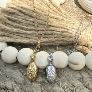 Peut inclure: Deux colliers avec des pendentifs ovales. L'un est dor&eacute; avec un motif floral, l'autre argent&eacute; avec un motif floral similaire. Les deux pendentifs sont suspendus &agrave; de fines cha&icirc;nes. Les colliers reposent sur une surface tiss&eacute;e.