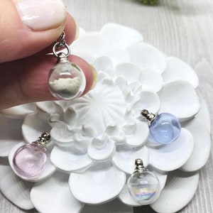 May include: Four glass bottle pendants with silver caps. The bottles are filled with different materials, including sand, iridescent glitter, and clear liquid. The pendants are displayed on a white flower-shaped surface.