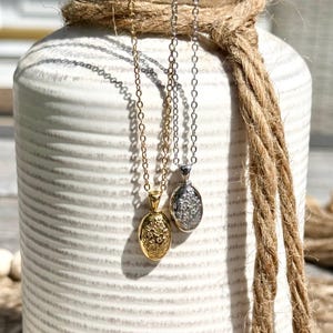 May include: Two necklaces with oval pendants hang in front of a white vase wrapped with rope. One necklace is gold-colored, the other is silver-colored. The pendants have floral designs.