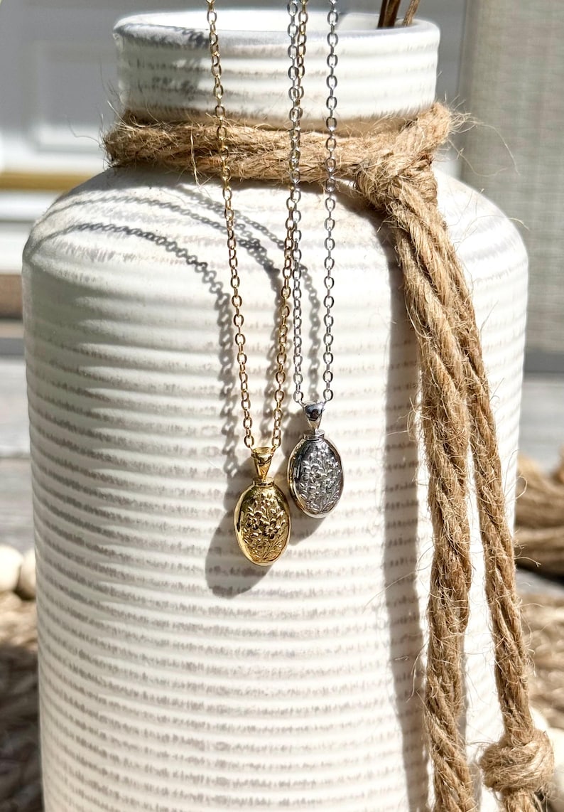 May include: Two necklaces with oval pendants hang in front of a white vase wrapped with rope. One necklace is gold-colored, the other is silver-colored. The pendants have floral designs.