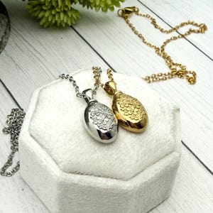 May include: Two oval-shaped pendant necklaces. One is silver-toned with a floral design, the other is gold-toned with a similar floral pattern. Both pendants hang on delicate chains. The necklaces rest on a white, textured surface.