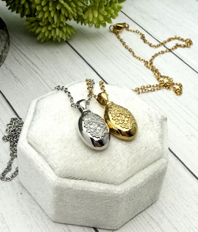 May include: Two oval-shaped pendant necklaces. One is silver-toned with a floral design, the other is gold-toned with a similar floral pattern. Both pendants hang on delicate chains. The necklaces rest on a white, textured surface.