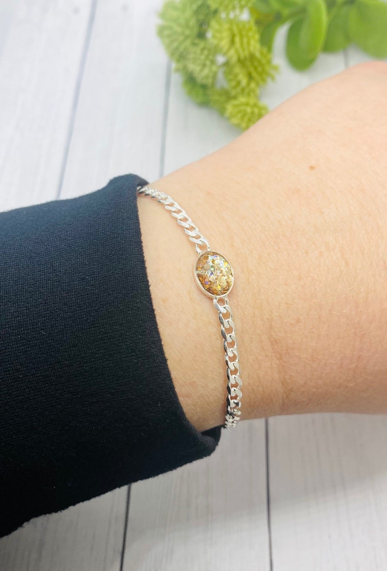May include: A silver-toned curb chain bracelet featuring a central oval-shaped charm. The charm contains a textured, multi-colored inlay with gold, bronze, and iridescent glitter flecks embedded within a smooth, rounded, metallic-rimmed setting.