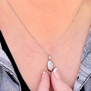 May include: A silver necklace with a small, oval pendant. The pendant is encrusted with small, sparkling stones. The necklace is displayed against a black and white plaid shirt.