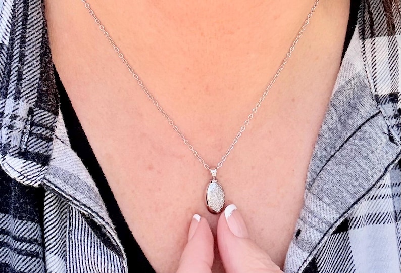 May include: A silver necklace with a small, oval pendant. The pendant is encrusted with small, sparkling stones. The necklace is displayed against a black and white plaid shirt.