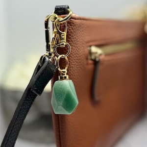 May include: A green gemstone charm with a gold-colored clasp is attached to a black leather strap. The charm is a faceted, geometric shape. The background features a brown leather purse with a gold zipper.