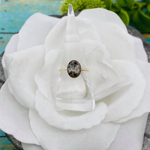 May include: A gold ring with a clear oval stone containing a pressed flower design. The ring is displayed on a white flower.