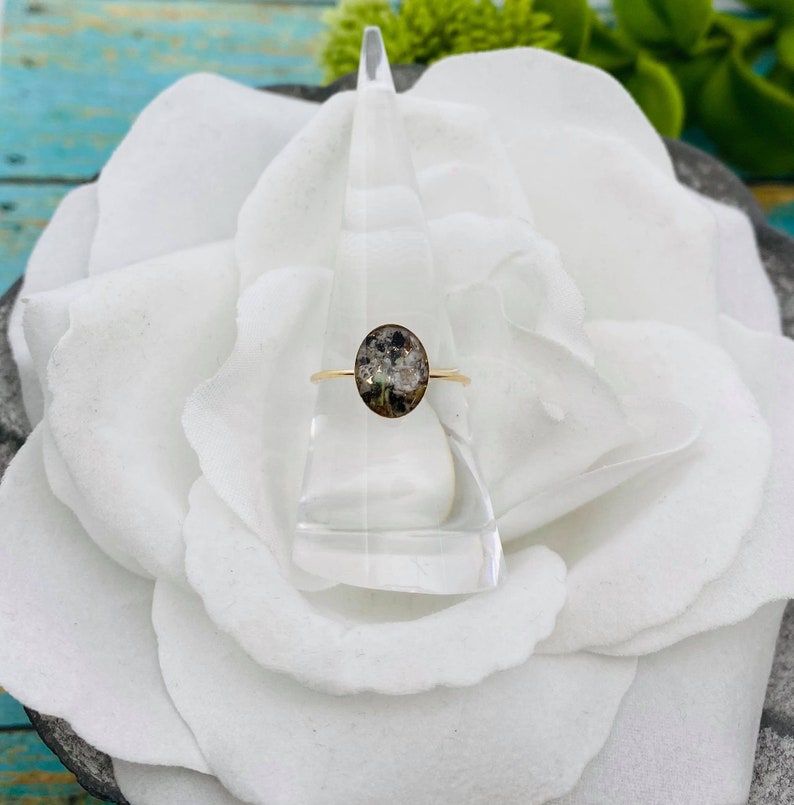 May include: A gold ring with a clear oval stone containing a pressed flower design. The ring is displayed on a white flower.