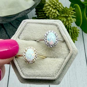 May include: Two oval opal rings with halo settings are displayed in a gray velvet ring box. One ring has a gold band, the other a silver band. The opals have a milky white base with flashes of color.