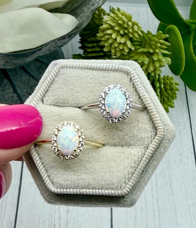 May include: Two oval opal rings with halo settings are displayed in a gray velvet ring box. One ring has a gold band, the other a silver band. The opals have a milky white base with flashes of color.