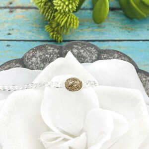 May include: A silver chain bracelet featuring a central oval-shaped charm with a textured, shimmering gold-toned surface. The bracelet is displayed resting on a white fabric surface, highlighting the contrast between the metallic silver chain and the gold center.