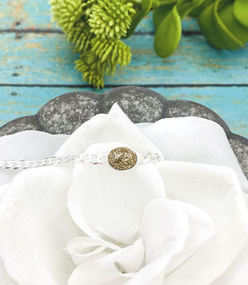 May include: A silver chain bracelet featuring a central oval-shaped charm with a textured, shimmering gold-toned surface. The bracelet is displayed resting on a white fabric surface, highlighting the contrast between the metallic silver chain and the gold center.