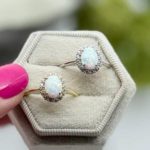 May include: Two opal rings with halo settings are displayed in a gray velvet ring box. One ring has a gold band, the other a silver band. The opals have a white base color with flashes of color.