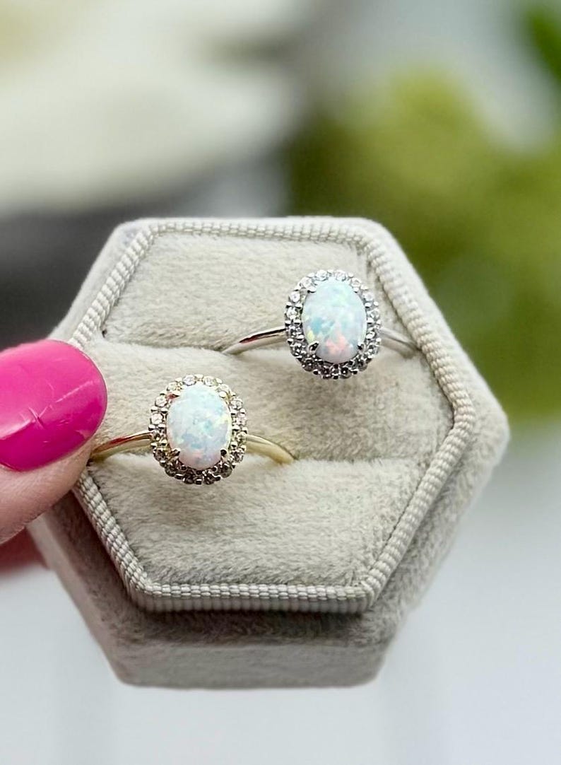 May include: Two opal rings with halo settings are displayed in a gray velvet ring box. One ring has a gold band, the other a silver band. The opals have a white base color with flashes of color.