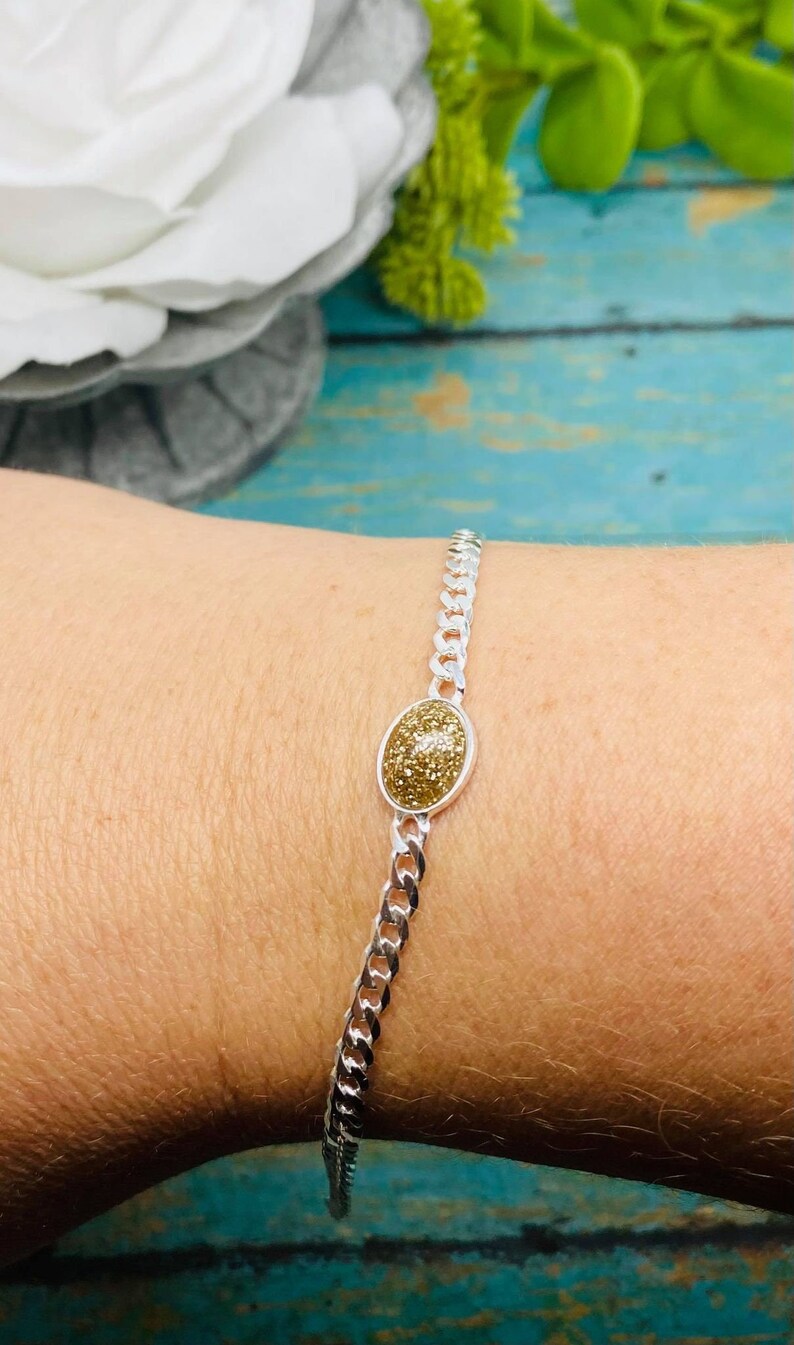 May include: A silver-toned chain bracelet featuring a central oval-shaped charm with a textured, shimmering gold-colored surface. The bracelet consists of interlocking metal links that form a continuous band around the wrist, showcasing a metallic finish.
