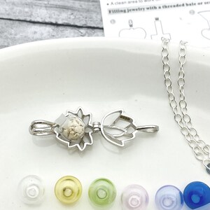 K&ouml;nnte beinhalten: Eine silberne Halskette mit einem Anh&auml;nger in Blumenform. Der Anh&auml;nger ist ge&ouml;ffnet und mit einer wei&szlig;en Substanz gef&uuml;llt. Die Halskette liegt auf einer wei&szlig;en Oberfl&auml;che mit einem kleinen Trichter, einem Zahnstocher und einer Kette.
