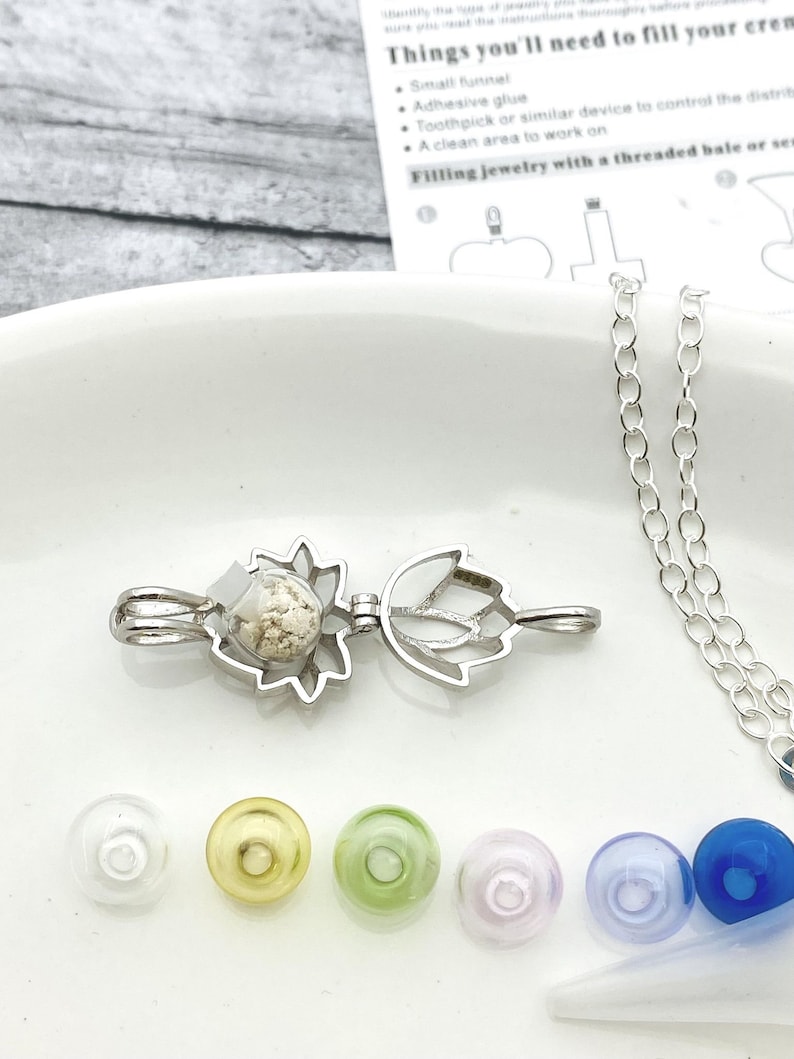 K&ouml;nnte beinhalten: Eine silberne Halskette mit einem Anh&auml;nger in Blumenform. Der Anh&auml;nger ist ge&ouml;ffnet und mit einer wei&szlig;en Substanz gef&uuml;llt. Die Halskette liegt auf einer wei&szlig;en Oberfl&auml;che mit einem kleinen Trichter, einem Zahnstocher und einer Kette.
