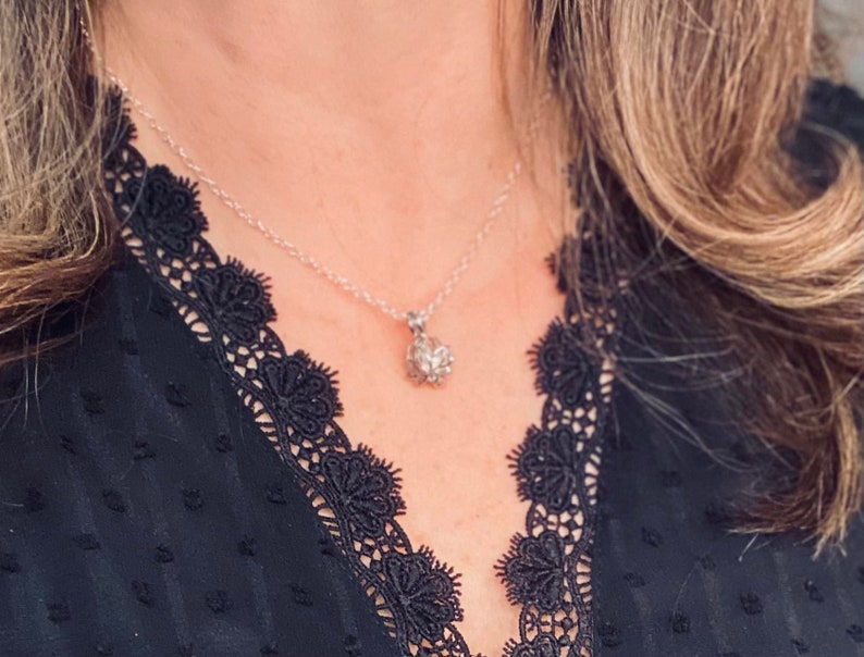 K&ouml;nnte beinhalten: Eine silberne Halskette mit einem kleinen, glitzernden Anh&auml;nger h&auml;ngt an einer Person. Die Kette wird &uuml;ber einem schwarzen Spitzenoberteil getragen.
