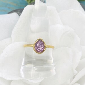 Może przedstawiać: Złoty pierścionek z fioletowym, błyszczącym kamieniem w kształcie łzy.