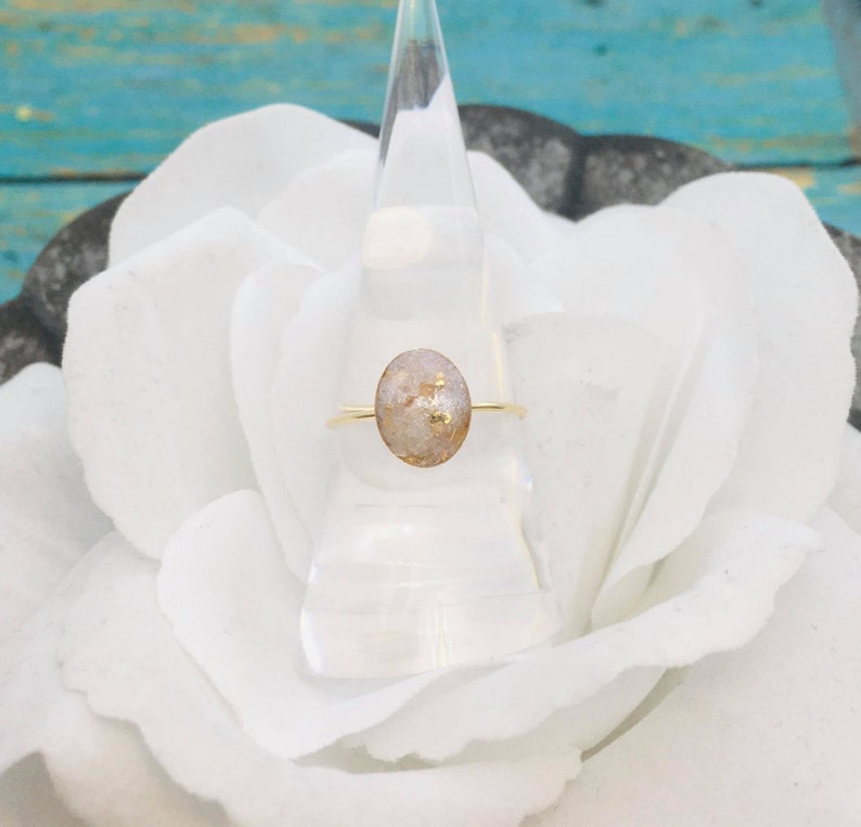 May include: A gold ring with a white oval-shaped stone embedded with gold flecks. The ring is displayed on a white flower.