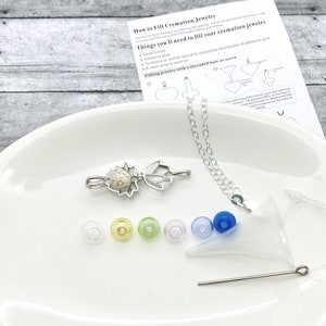 K&ouml;nnte beinhalten: Ein silbernes Krematoriums-Schmuckset mit einem Lotusbl&uuml;ten-Anh&auml;nger, einem kleinen Trichter, einem Zahnstocher und verschiedenen farbigen Stopfen. Das Set enth&auml;lt Anweisungen zum Bef&uuml;llen des Schmucks mit einem Gewinde-B&uuml;gel oder einer Schraube.
