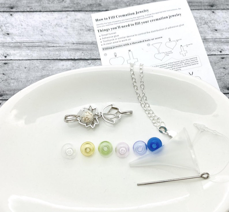 K&ouml;nnte beinhalten: Ein silbernes Krematoriums-Schmuckset mit einem Lotusbl&uuml;ten-Anh&auml;nger, einem kleinen Trichter, einem Zahnstocher und verschiedenen farbigen Stopfen. Das Set enth&auml;lt Anweisungen zum Bef&uuml;llen des Schmucks mit einem Gewinde-B&uuml;gel oder einer Schraube.