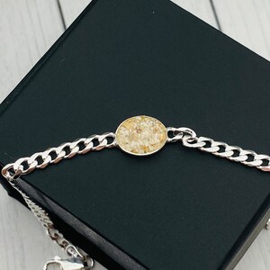May include: A silver-toned curb chain bracelet featuring a central circular pendant. The pendant contains a textured inlay of small, light beige and cream-colored granular pieces set within a smooth, polished metal frame that matches the chain.