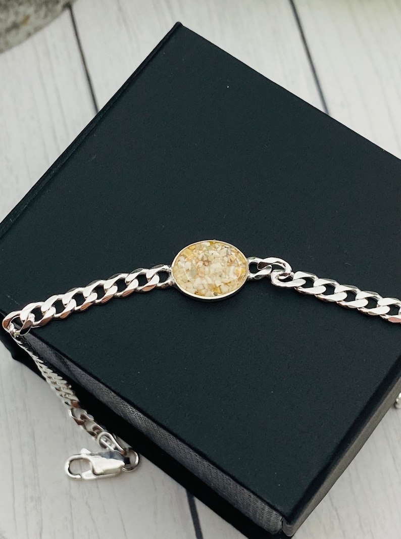 May include: A silver-toned curb chain bracelet featuring a central circular pendant. The pendant contains a textured inlay of small, light beige and cream-colored granular pieces set within a smooth, polished metal frame that matches the chain.
