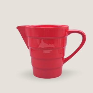 May include: A bright red ceramic pitcher with a handle and a spout. The pitcher has a series of horizontal ridges around its body. The handle is curved and attached to the side of the pitcher. The spout is triangular and angled. The pitcher is a solid, glossy red.