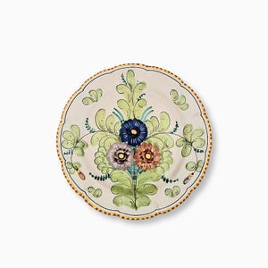 Könnte beinhalten: Ein runder Keramikteller mit gewelltem Rand und handgemaltem Blumenmuster. In der Mitte befinden sich drei stilisierte Blüten in Blau, Lavendel und Braun, umgeben von hellgrünen Blattornamenten und einem Rand mit gelben Punkten.