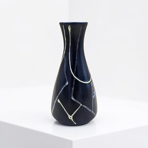 Könnte beinhalten: Eine schwarze Keramikvase mit schmalem Hals und bauchigem Körper. Die Vase ist mit weißen und hellgelben abstrakten Linien verziert. Die Vase wird auf einer weißen Oberfläche präsentiert.