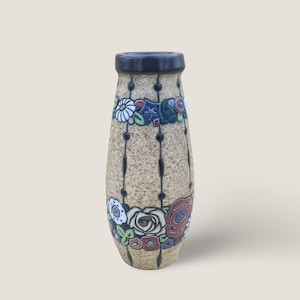 Op de afbeelding: Een decoratieve keramische vaas met een gespikkelde bruine body en een donkerblauwe rand. De vaas heeft bloemen en geometrische ontwerpen in rood, wit, blauw en groen. Zwarte lijnen met kleine zwarte stippen lopen verticaal.