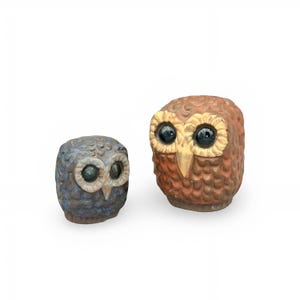 Puede incluir: Dos figuras de búho de cerámica con superficies texturizadas que imitan plumas. El búho más pequeño es azul con la cara clara, mientras que el más grande es marrón con la cara color crema. Ambos tienen ojos grandes, oscuros y brillantes.