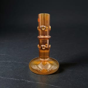 Könnte beinhalten: Eine handgefertigte, bernsteinfarbene Glasvase mit rundem Boden und zylindrischem Oberteil. Die Vase hat dekorative kreisförmige Akzente und eine glatte, reflektierende Oberfläche. Das Design erinnert an Vintage-Glaswaren.