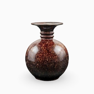 Può includere: Un vaso in ceramica rotondo con finitura lucida, caratterizzato da uno smalto maculato marrone scuro e nero. Il vaso presenta un collo stretto con tre nervature orizzontali e un bordo largo, piatto e svasato con un corpo bulboso.