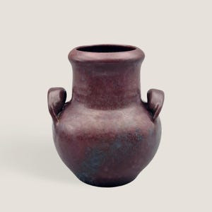 Könnte beinhalten: Eine maroonfarbene Keramikvase mit zwei gebogenen Griffen. Die Vase hat einen breiten, abgerundeten Körper, der sich zur Basis hin verjüngt, und einen ausgestellten Rand. Die Oberfläche hat ein gesprenkeltes Aussehen mit dunkleren Bereichen, was auf eine strukturierte Oberfläche hindeutet.