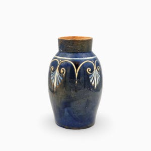 May include: A ceramic vase with a deep blue glazed finish, featuring a tan rim and a decorative white and light blue painted scroll pattern around the upper shoulder. The body of the vase has a smooth, glossy texture with subtle colour variations.