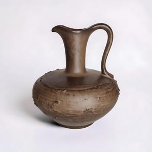 Könnte beinhalten: Braune Keramikkrug mit rundem Körper und gebogenem Henkel. Der Krug hat einen Ausguss und eine strukturierte Oberfläche mit kleinen, erhabenen Noppen. Das Design ist schlicht und elegant, geeignet zum Servieren von Getränken oder als dekoratives Element.