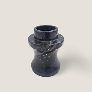 Könnte beinhalten: Ein Flaschenverschluss aus schwarzem Marmor mit zylindrischem Oberteil und bauchiger Basis. Der Verschluss hat eine polierte Oberfläche mit weißen Adern. Das Design ist eine klassische Form für eine Weinflasche.