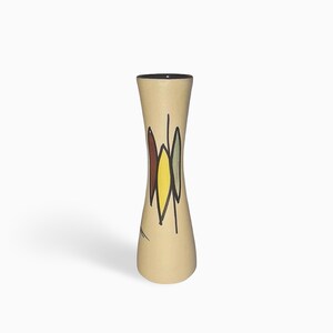 Könnte beinhalten: Eine hohe, sanduhrförmige Keramikvase mit matter, cremefarbener Oberfläche. Das Äußere zeigt ein handgemaltes, abstraktes Design aus drei vertikalen, blattförmigen Motiven in Dunkelbraun, Gelb und Salbeigrün, die mit schwarzen Linien umrandet sind.